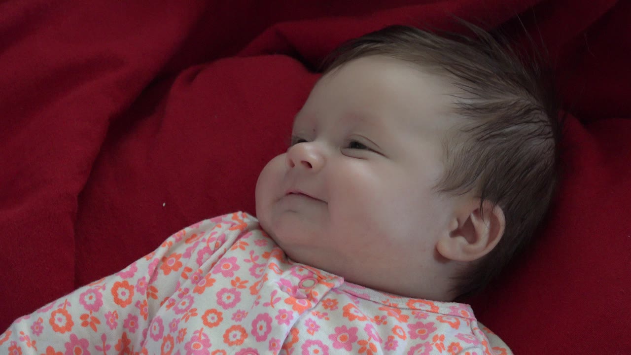Newborn baby lying on red bedding in floral pajamas.
