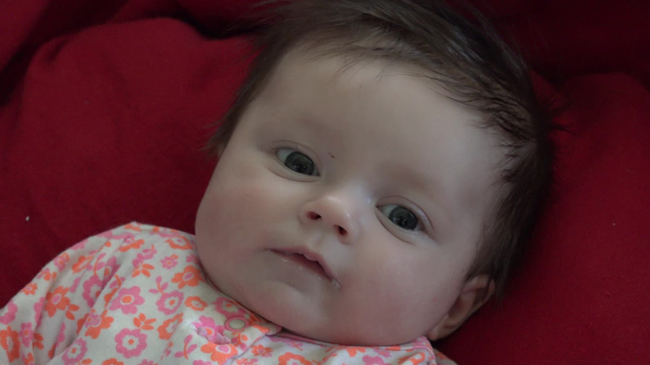 At 0:01, the baby remains reclined on the red blanket in Westminster, Colorado, with the face centered and floral clothing visible at the lower edge.
