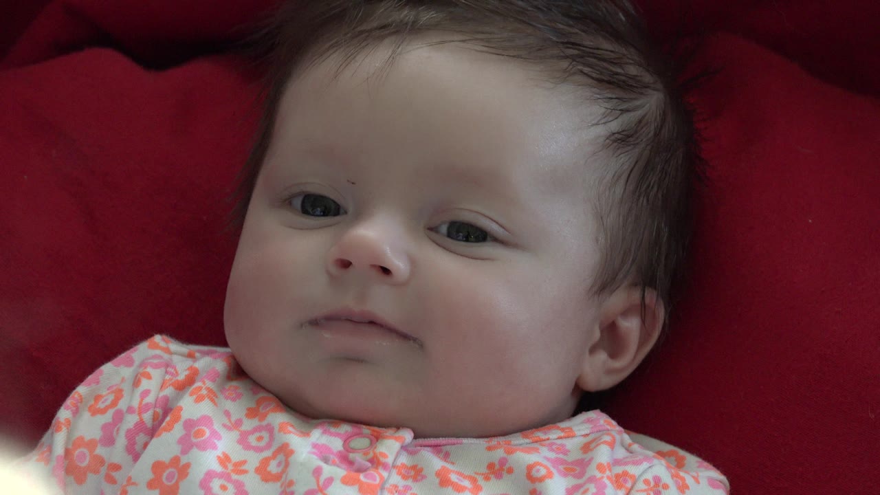 At 0:01, the baby's face remains centered against the red bedding in Westminster, Colorado, with eyes open and the floral clothing visible along the lower edge.