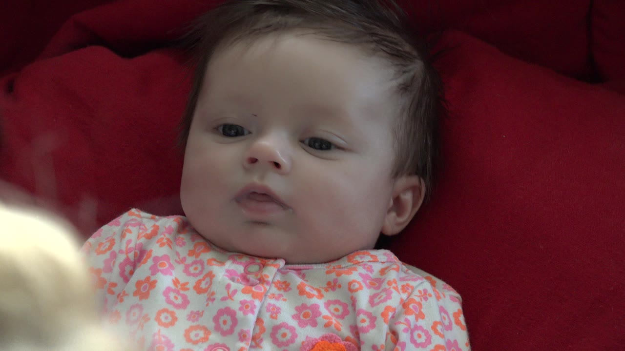 The baby remains on the red fabric in Westminster, Colorado, eyes open and mouth relaxed while a pale blurred foreground shape sits at the lower left edge.