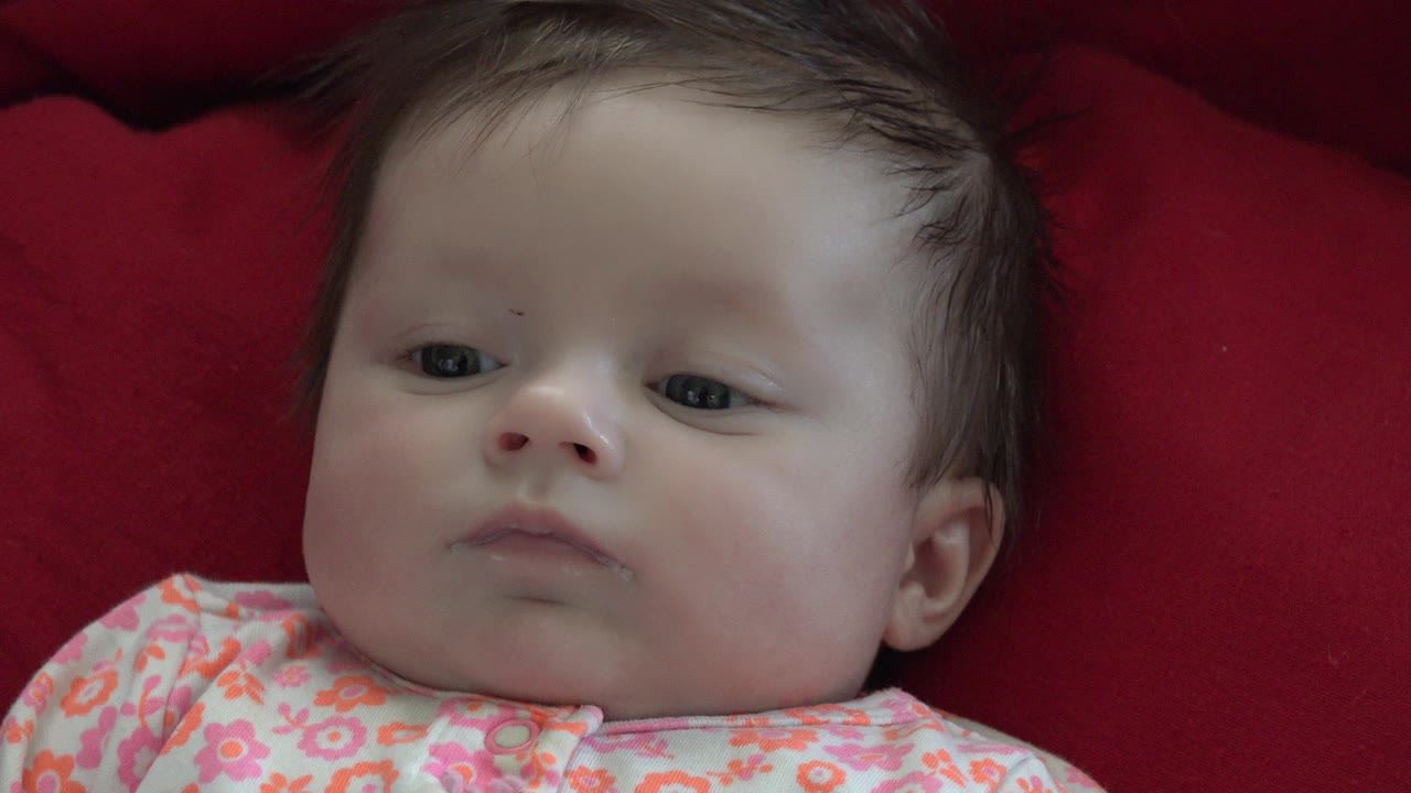 At 0:00, a baby lies on a deep red blanket in Westminster, Colorado, with the face angled slightly left and a pink floral top visible at the bottom of frame.
