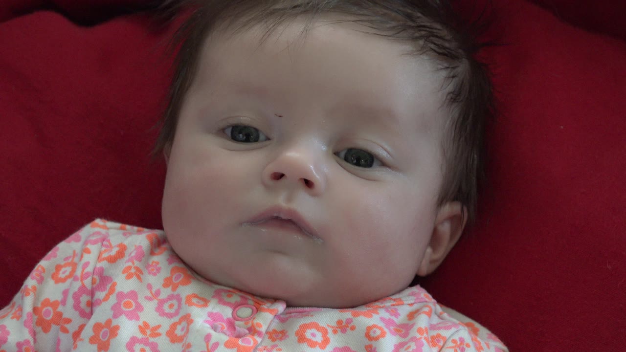 At 0:00, a dark-haired baby lies on red bedding in Westminster, Colorado, looking toward the camera with a neutral expression and floral clothing visible.