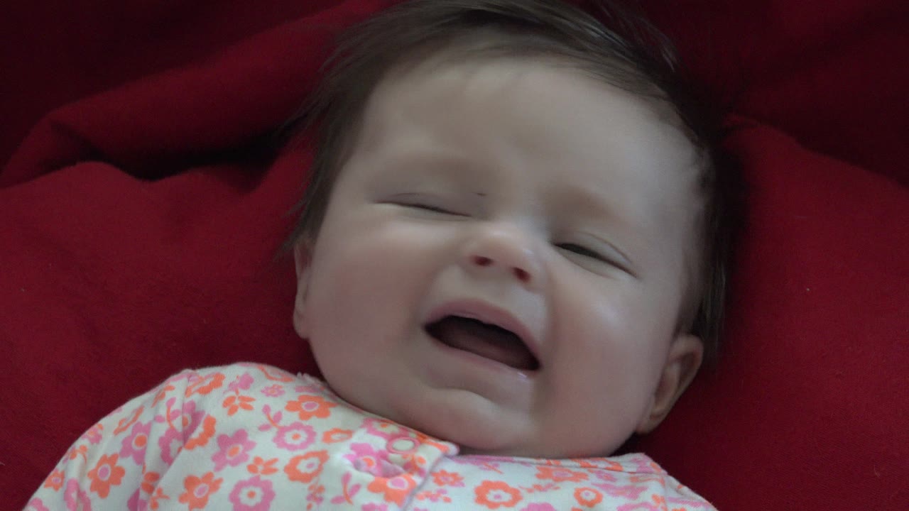 Baby lying on red fabric with mouth open and eyes nearly closed.