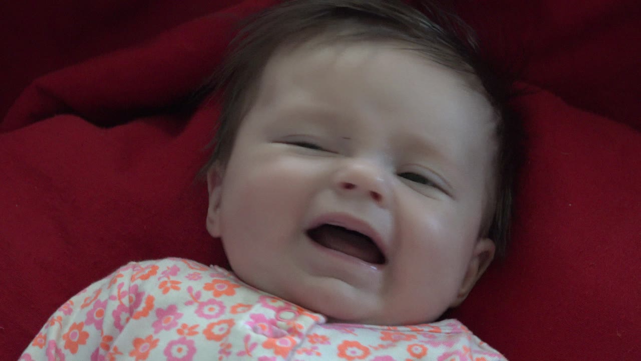 At 0:01 in Westminster, Colorado footage, the baby remains on the red fabric with one eye partly open and mouth still open, softening the crying expression.
