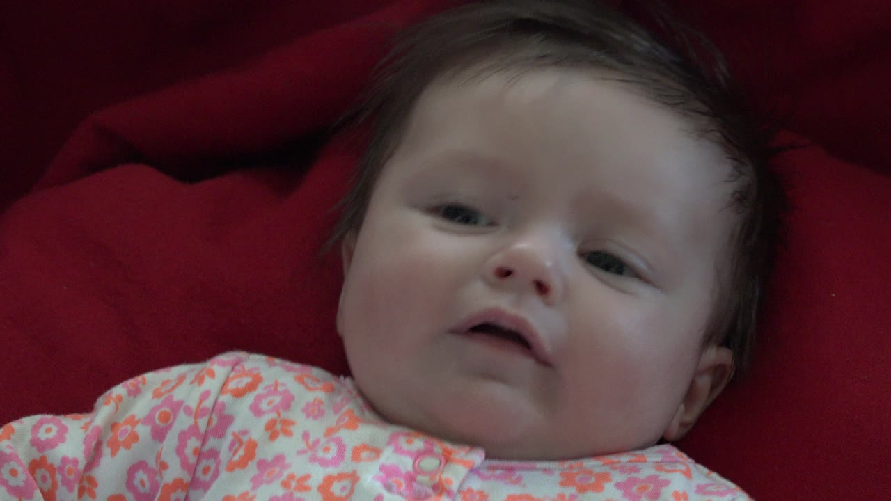 At 0:01, the baby remains reclined on red fabric, looking toward the camera with a slightly open mouth and soft light across the forehead and cheeks.