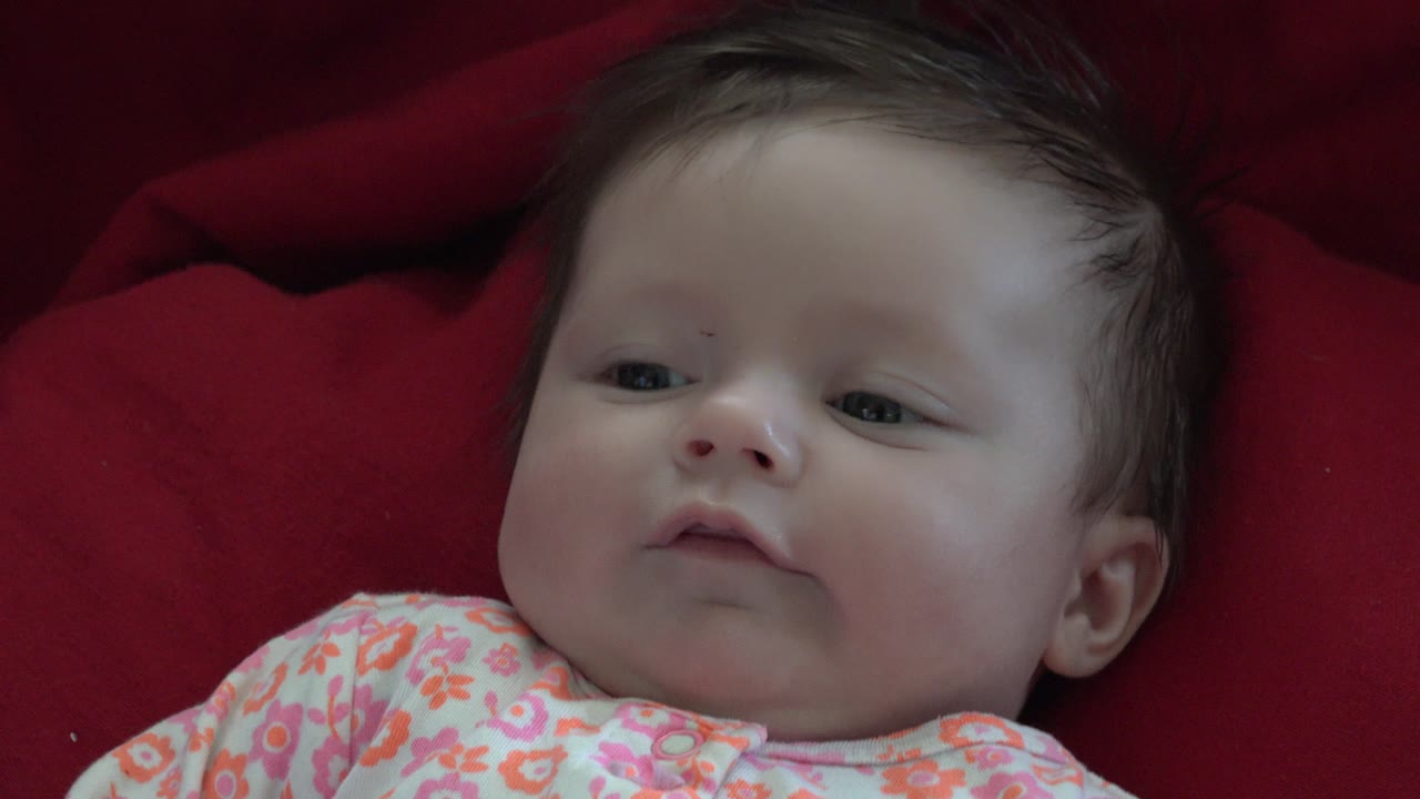 At 0:02, the baby's head angles slightly to the side, with the floral outfit lower in frame and the red fabric continuing as the plain background.