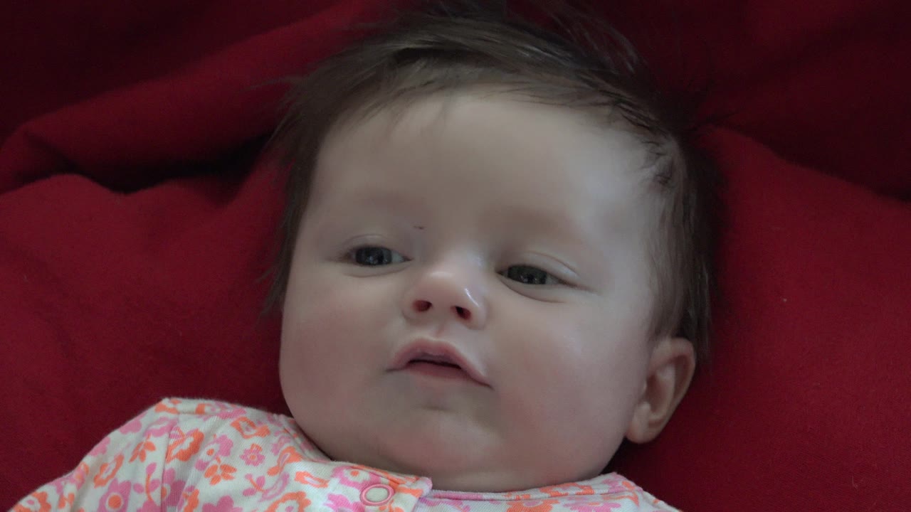 The baby remains close-framed on red fabric in Westminster, Colorado, with the mouth slightly parted and the eyes looking off to one side.