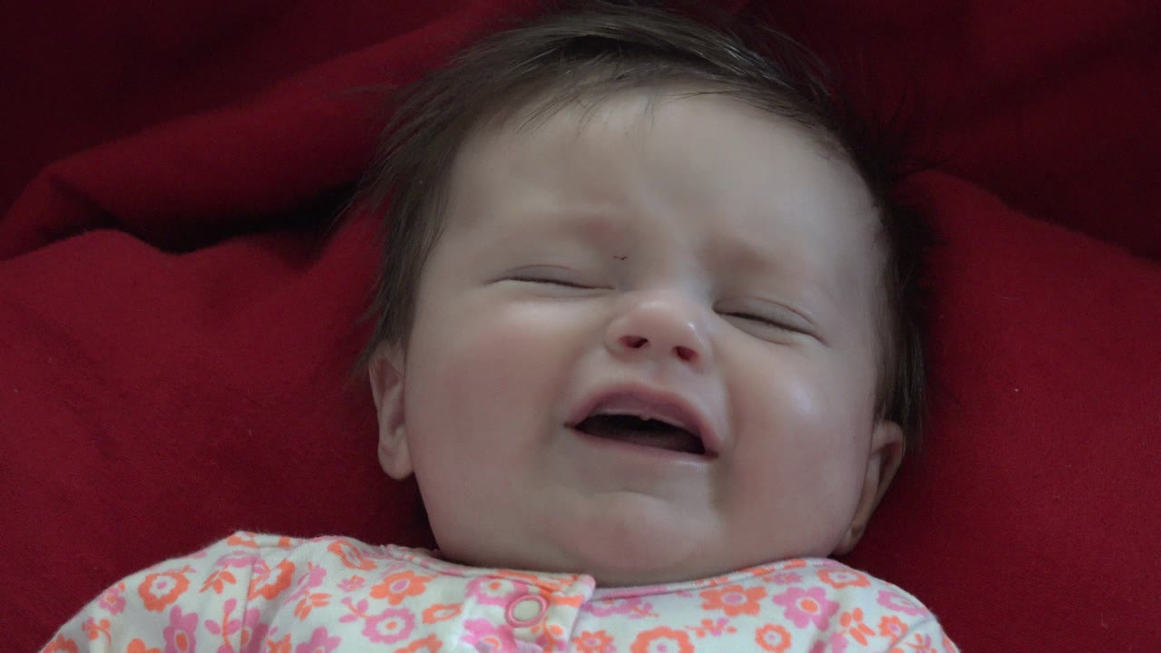 Baby lying on a red blanket with eyes closed and mouth open.