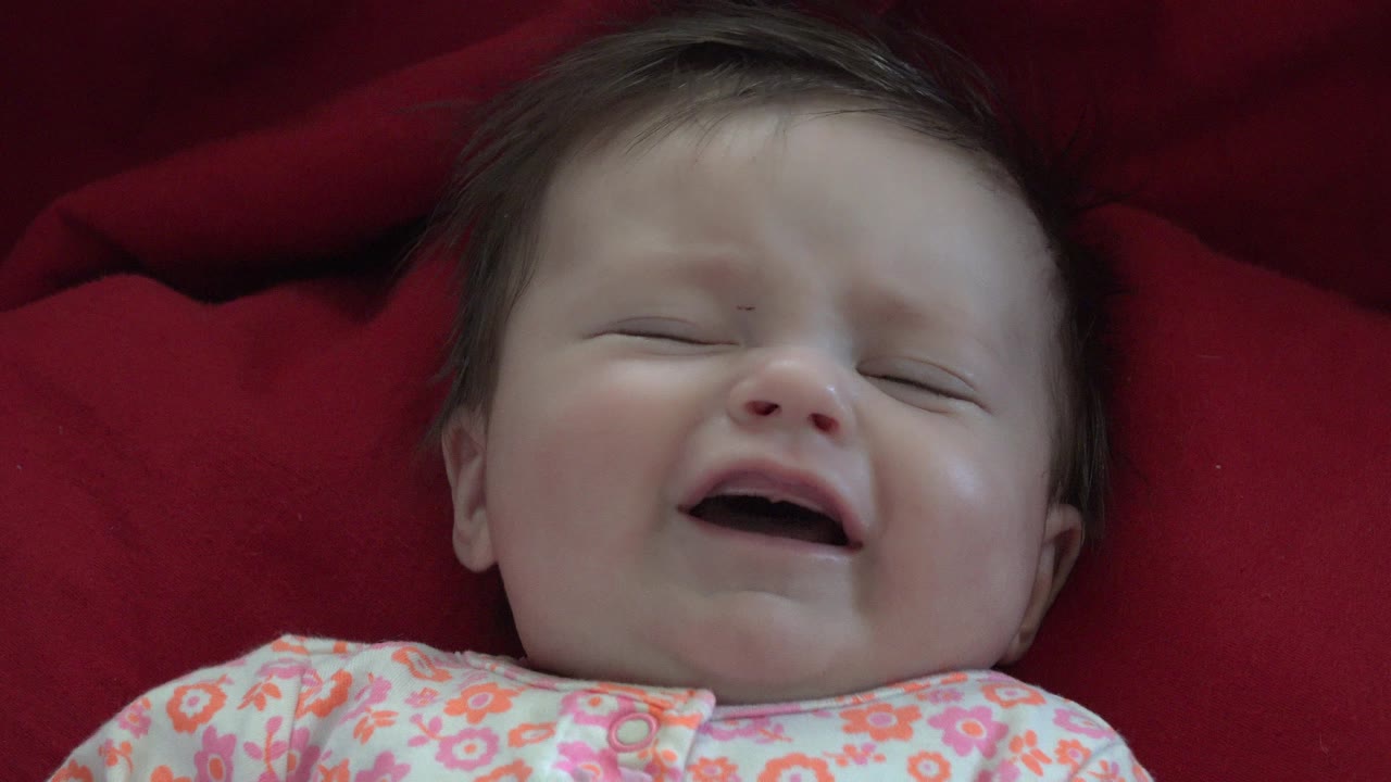 At 0:01 in Westminster, Colorado, the baby remains centered on the red blanket, smiling with eyes nearly closed and a floral onesie visible at the bottom of frame.