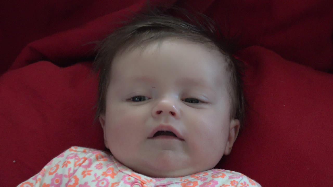 At 0:01, the baby remains centered on red pillows in Westminster, Colorado, with eyes partly open and the floral outfit visible at the bottom of frame.