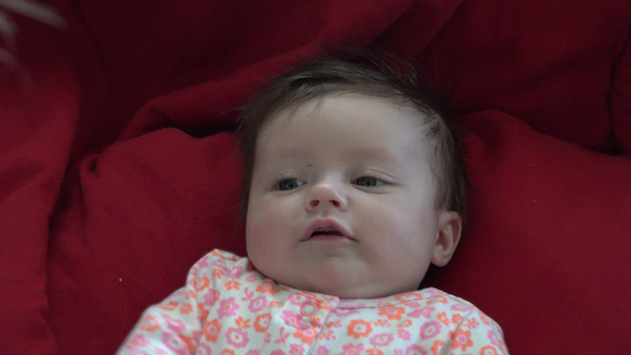 At 0:00, an infant in Westminster, Colorado lies low in frame on red cushions, wearing a pink and orange floral onesie and looking slightly off to the side.