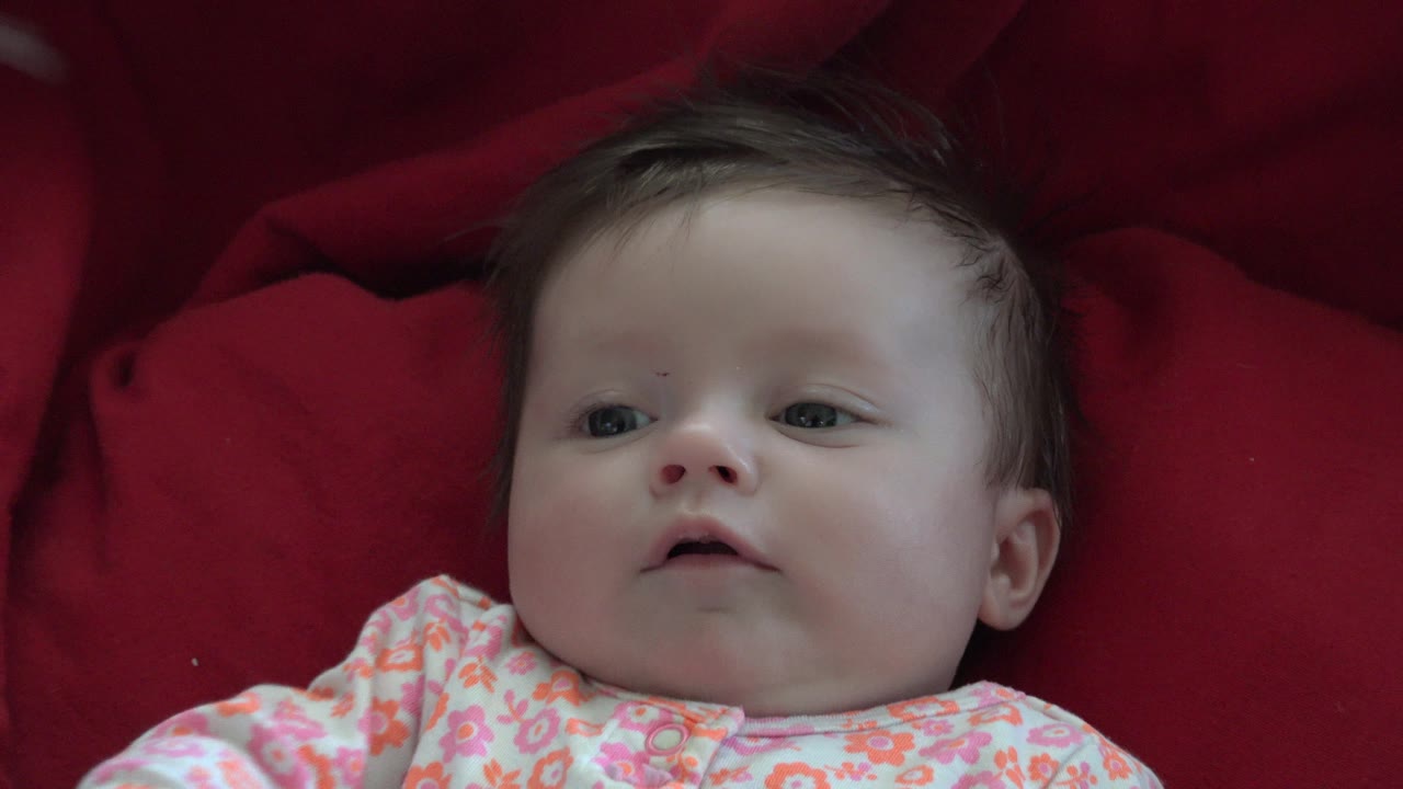 At 0:02, the infant's face is closer to the right side of the frame in Westminster, Colorado, with dark hair visible against the red fabric.