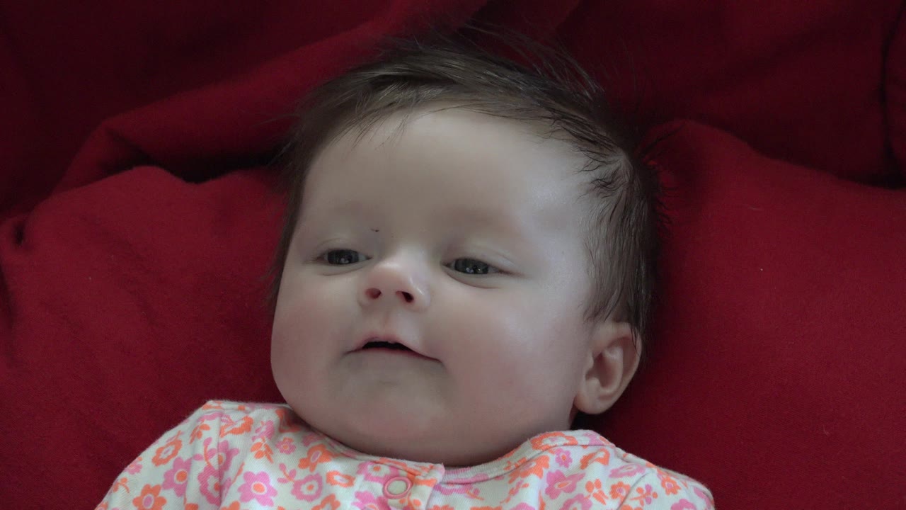 In Westminster, Colorado, a dark-haired baby lies on red bedding with the face angled slightly upward at the start of the clip.