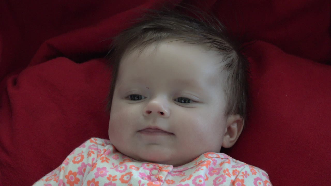 At 0:00, a baby in Westminster, Colorado lies close to camera on deep red fabric, wearing a white floral top with the face turned slightly left.