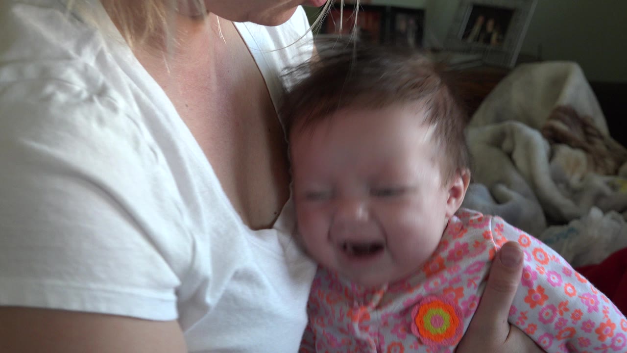 In Westminster, Colorado, an adult in a white shirt holds a crying baby in a pink floral outfit close to the camera inside a home.