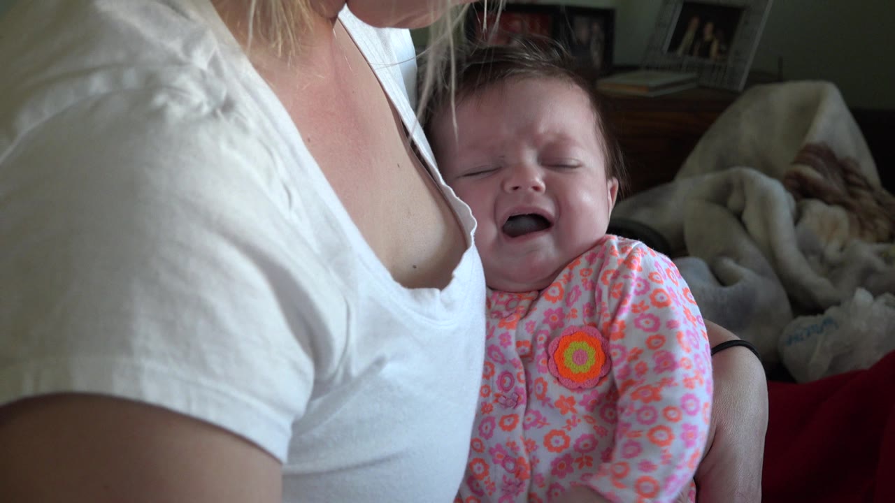 Crying baby held against an adult in a white shirt indoors.