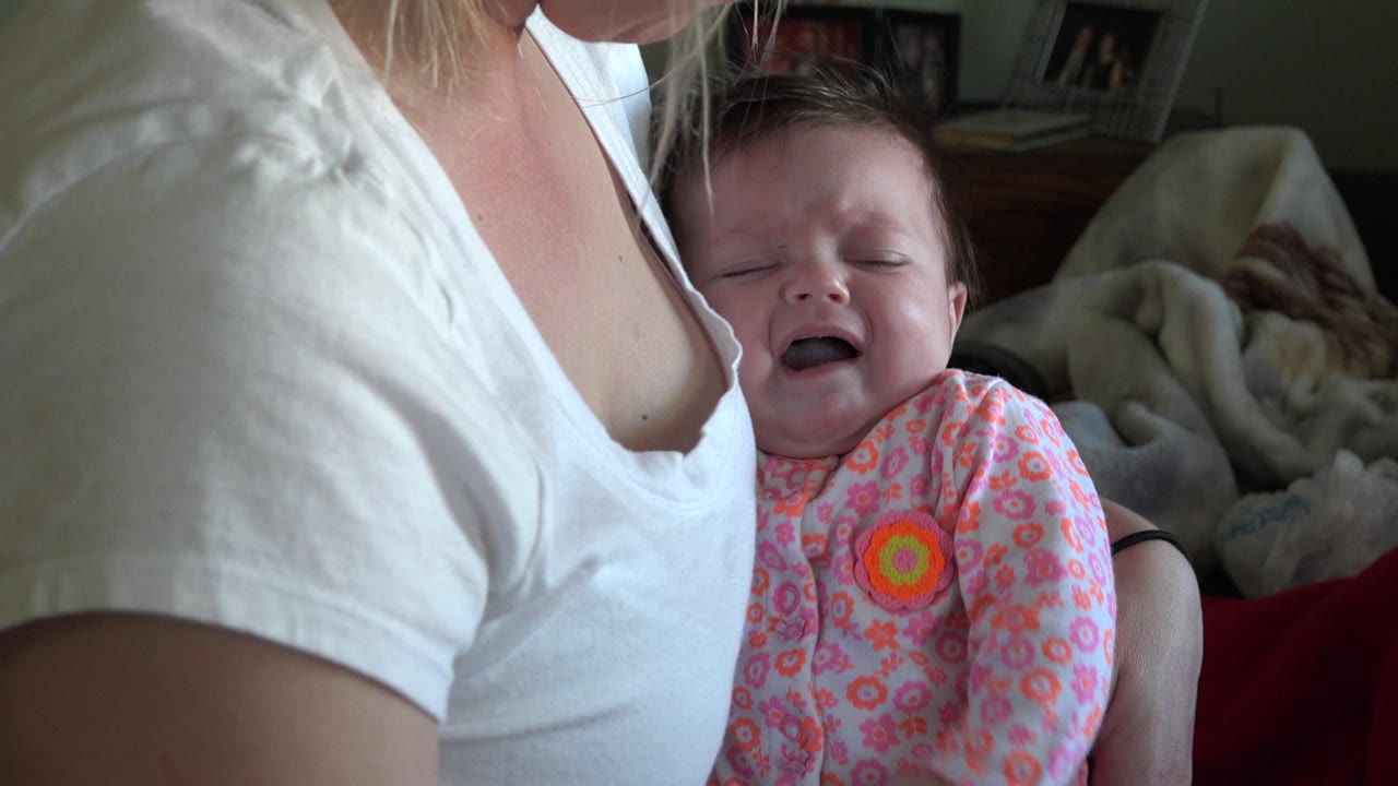 At 0:01 in Westminster, Colorado, the infant remains crying with eyes closed while pressed near the adult's shoulder, with soft bedding behind them.