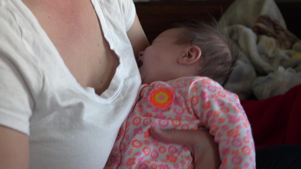 In Westminster, Colorado, a small infant in a pink floral onesie rests against a woman in a white V-neck shirt in a dim indoor setting.