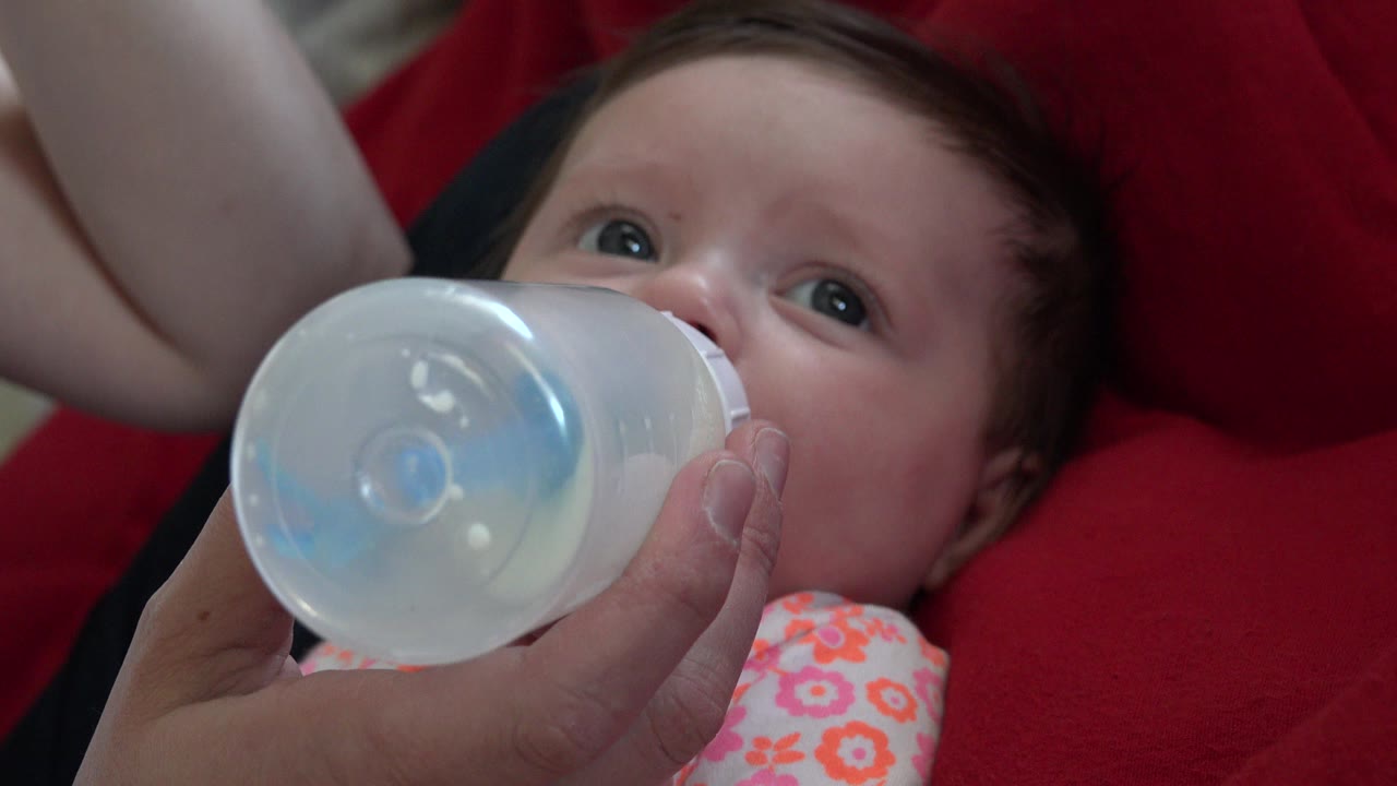 At the start of the Westminster, Colorado clip, a baby lies on red bedding while an adult hand holds a translucent milk bottle over the baby's mouth.