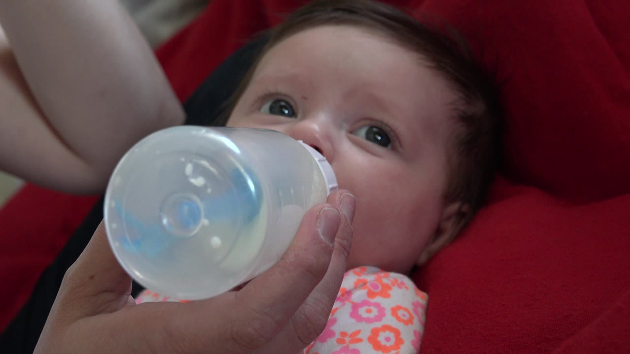 One second into the Westminster, Colorado footage, the baby's eyes look upward as the bottle stays pressed near the mouth and red fabric fills the background.