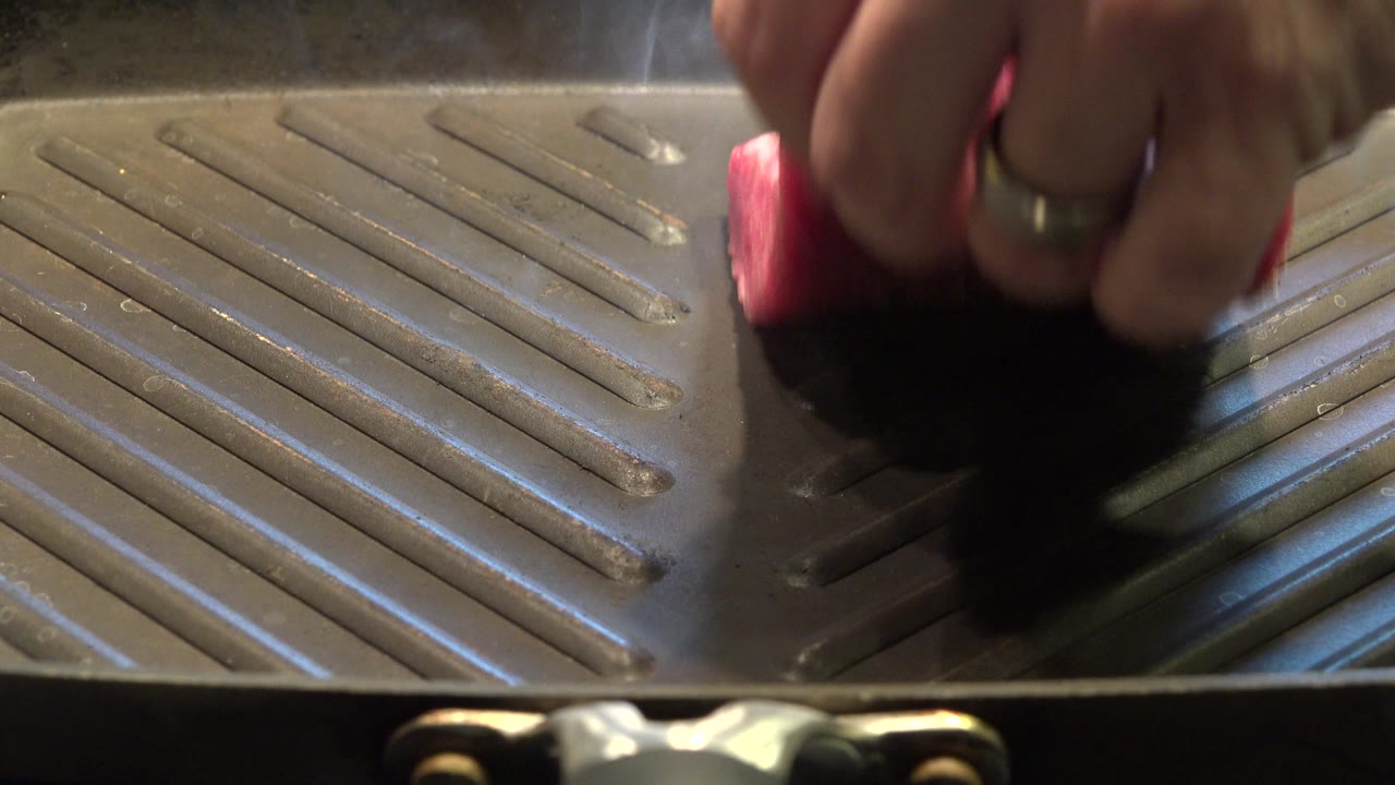 A partial hand lowers a piece of raw red meat onto a dark ridged grill pan in Westminster, Colorado, with warm light and no visible face.