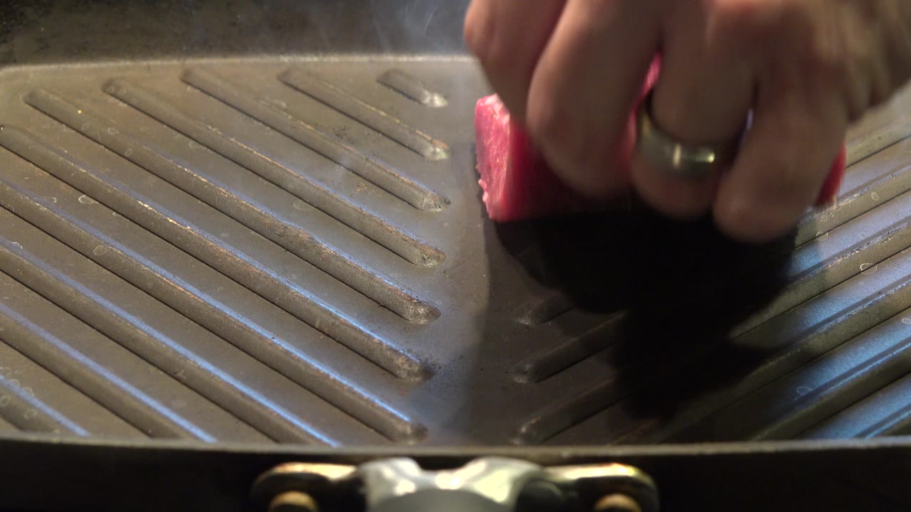 A partial hand presses or positions raw meat on the ridged pan surface in Westminster, Colorado, while warm highlights catch the metal and meat.