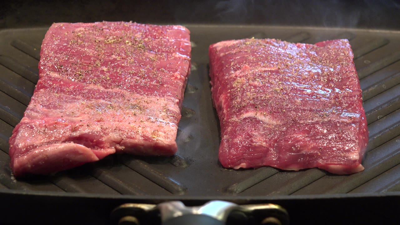 Two raw steaks on a black ridged grill pan with seasoning on top.