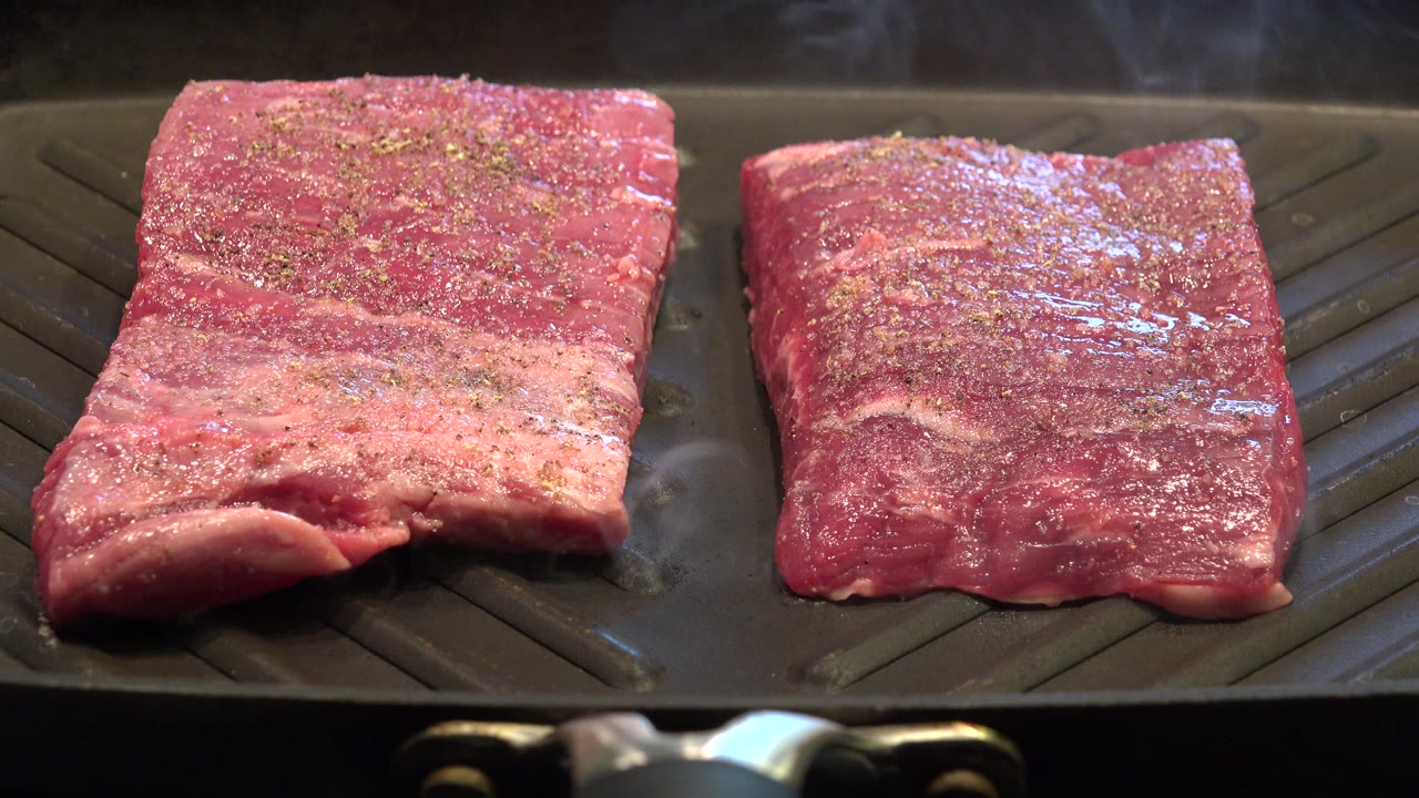 At 0:01, the steaks remain centered on the ridged pan, their wet red surfaces catching warm light while steam rises behind them and no visible people appear.