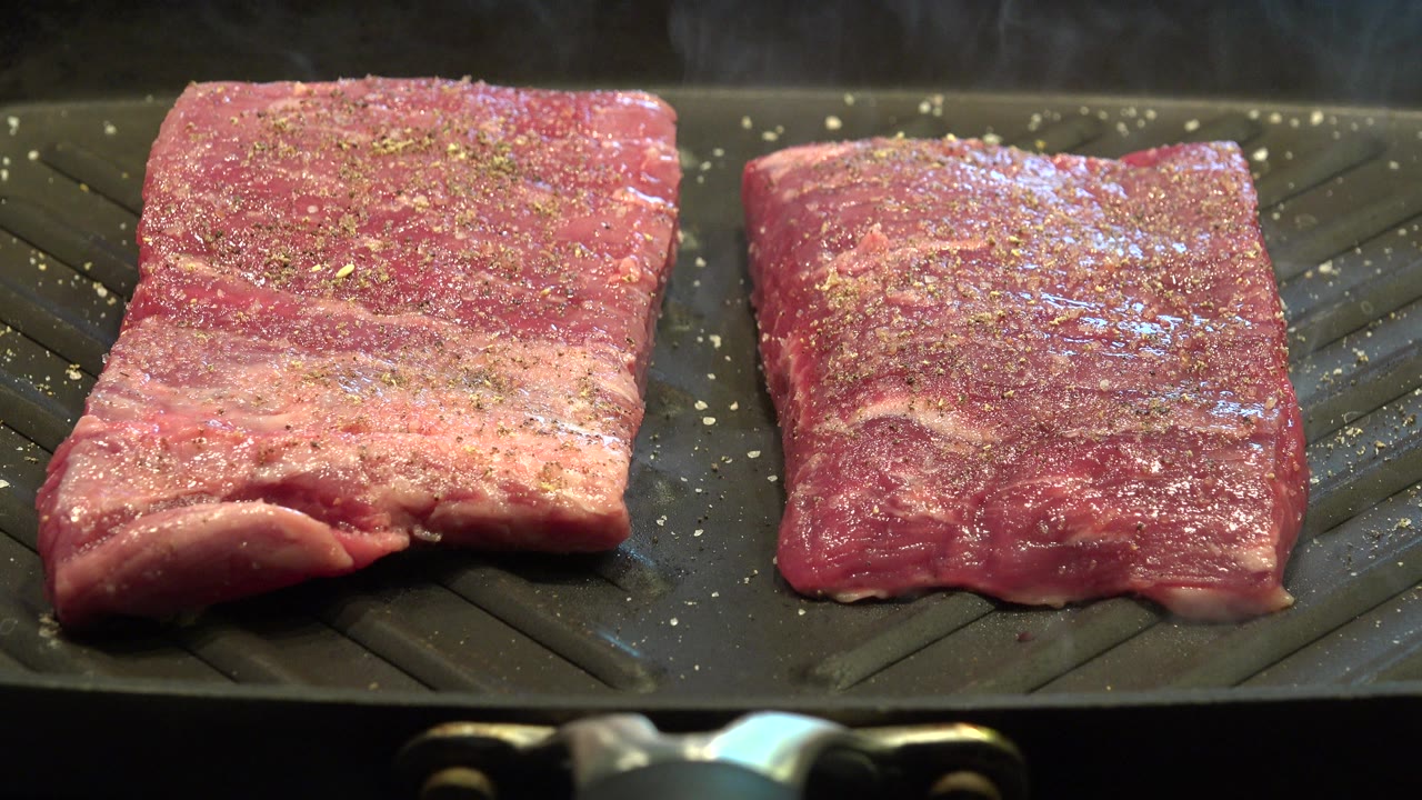 Two thick seasoned pieces of bacon lie side by side on a dark ridged grill pan in Westminster, Colorado, with glossy oil highlights and no visible people.
