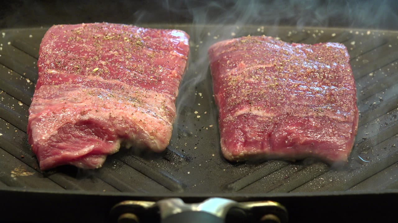 Two seasoned pieces of beef begin searing on a dark ridged grill pan in Westminster, Colorado, with light steam rising and no visible people.