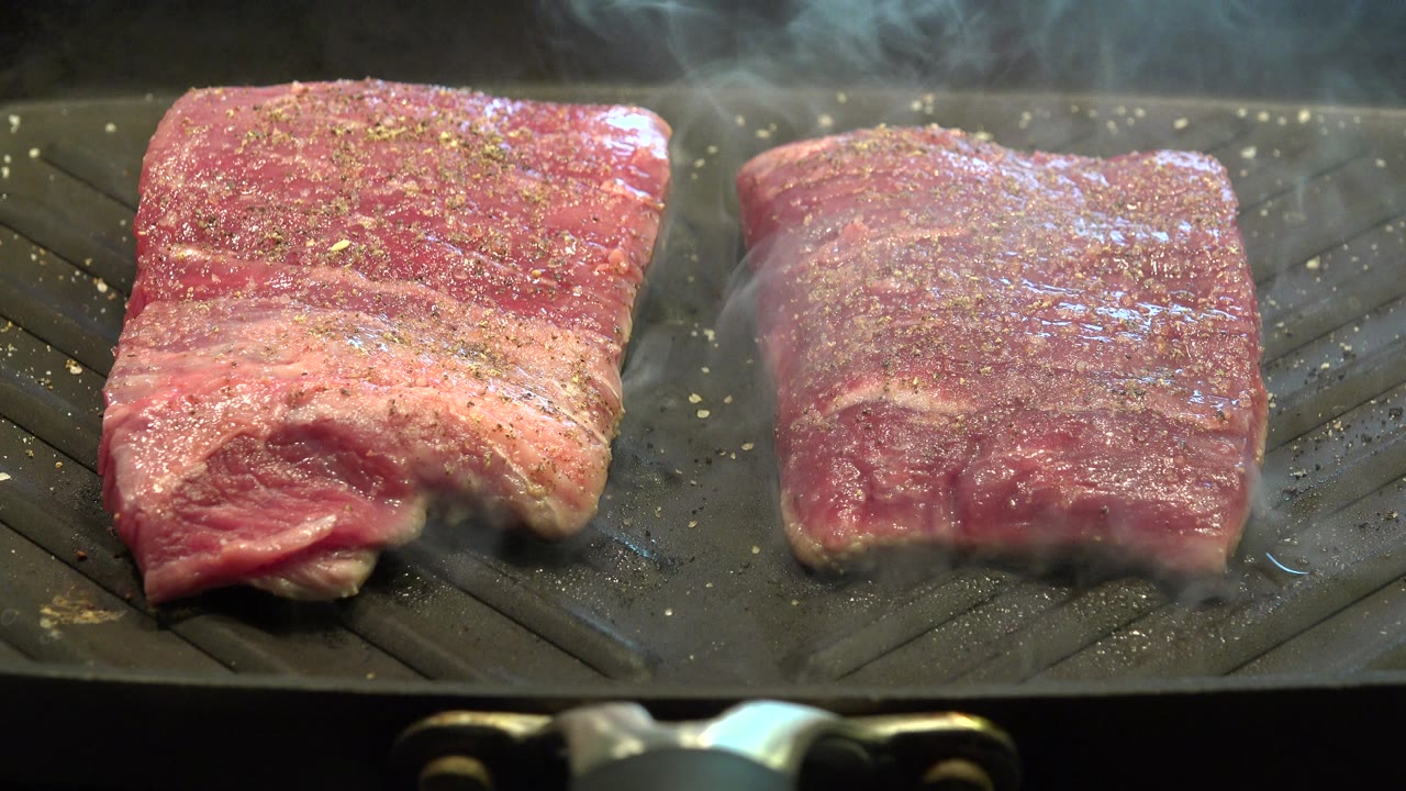 Two seasoned steaks on a smoky black ridged grill.