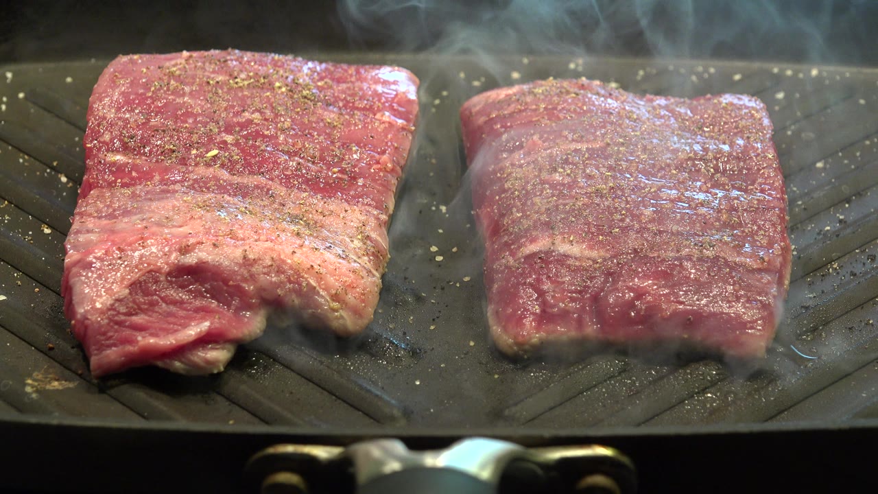 One second into the Westminster, Colorado footage, smoke drifts above two seasoned steak pieces on the black grill surface, with no visible people.
