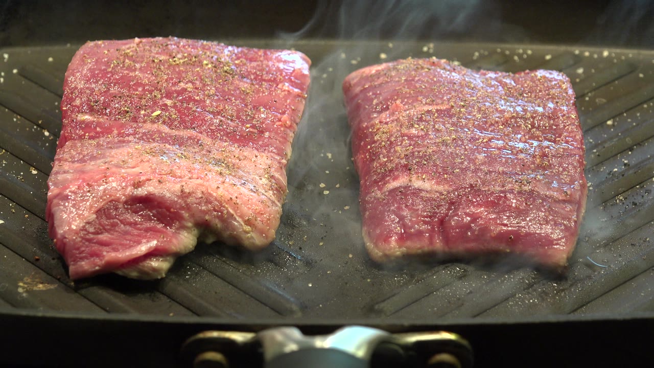 The steaks continue searing on the gray ridged grill, with moisture shining on the meat and no visible people in the frame.