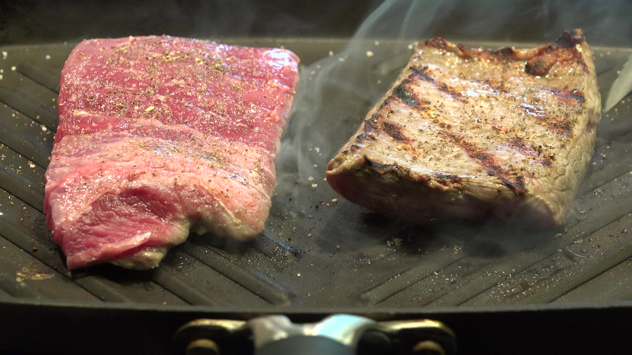 Two pieces of meat continue to cook as smoke rises from the grill pan.
