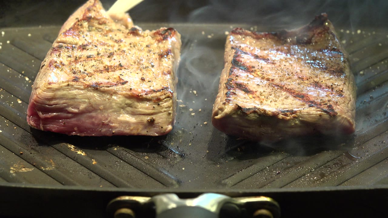At 0:00 in Westminster, Colorado, two thick pieces of food sit on a black ridged grill pan with browned sear marks, warm highlights, and no visible people.