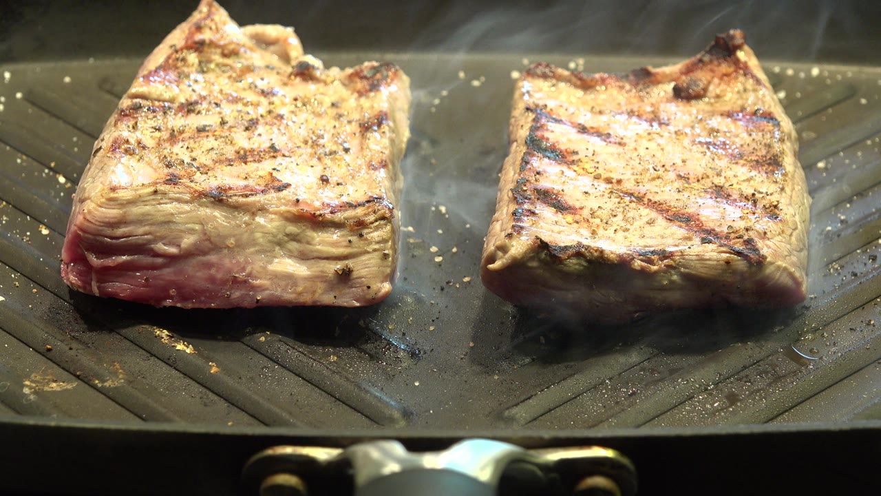 Two browned food pieces searing on a grill pan in warm light.