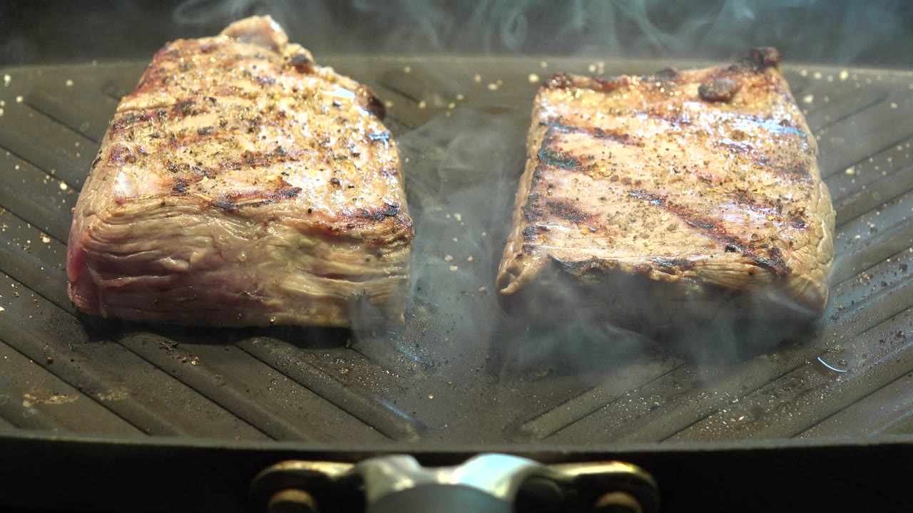 Two browned, seasoned meat pieces remain close together on the ridged grill in Westminster, Colorado, as a thicker band of smoke drifts across the upper edge with no visible people.