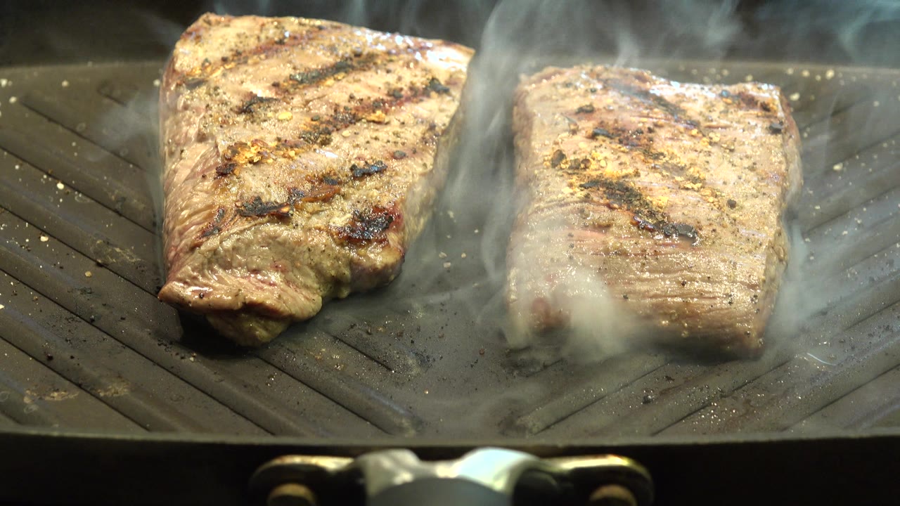 A close view of two steaks on the ridged pan shows heavier smoke drifting over the right piece, with no visible people.
