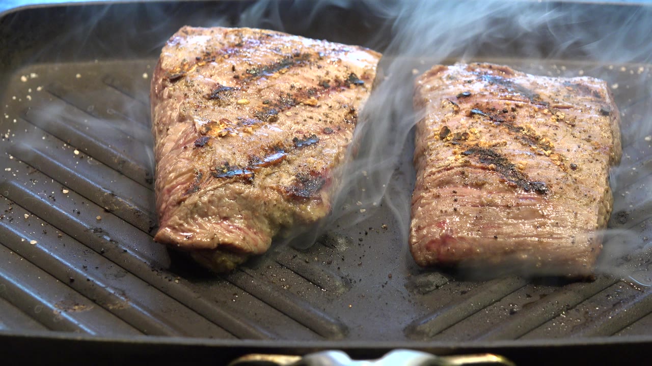 Two browned pieces of meat cook on a ridged fryer pan in Westminster, Colorado, with smoke drifting up and no visible people.