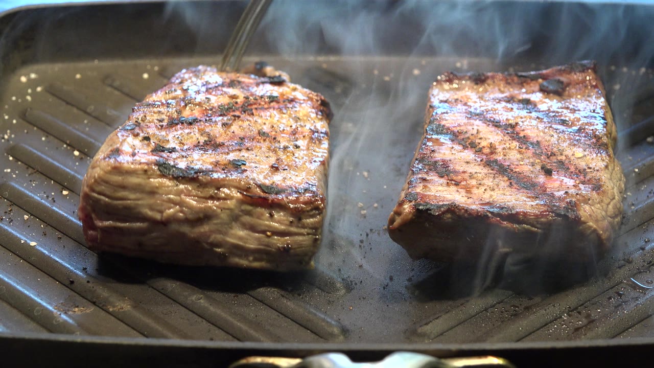 A fork or utensil appears near the upper edge as two pieces of grilled meat continue steaming on a ridged pan in Westminster, Colorado.
