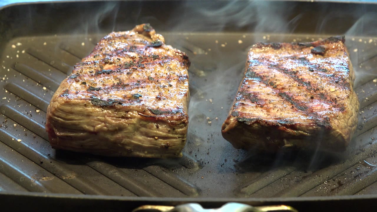 At 0:01, the same two meat pieces remain on the grill pan, with diagonal char marks more apparent on the right cut and no visible people.