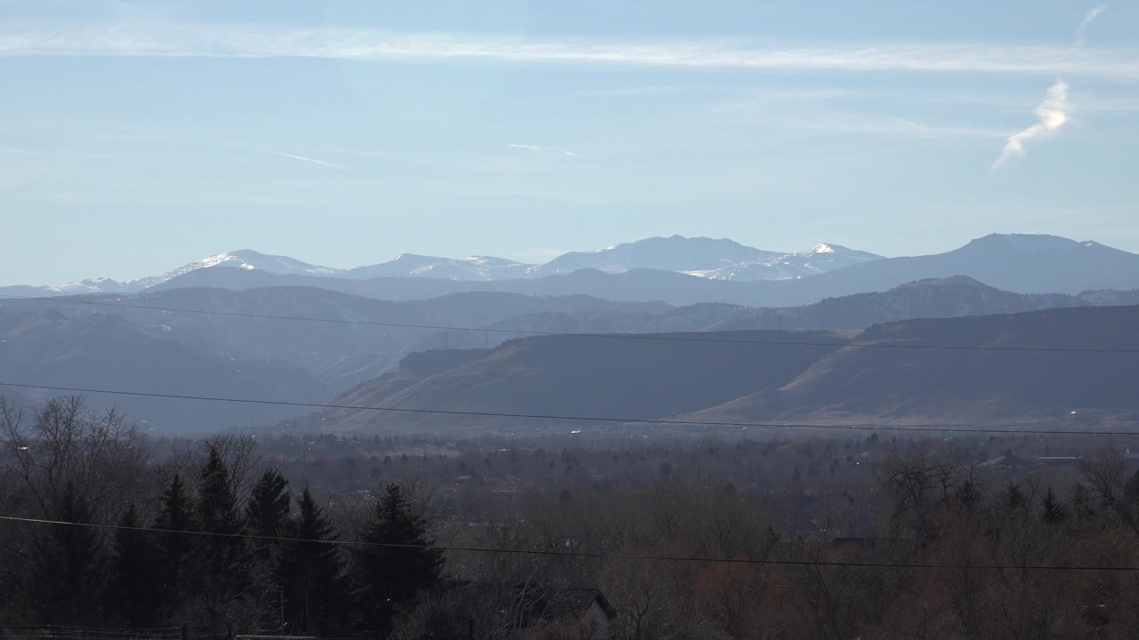 Arvada, Colorado mountain view with a broad dark hill band, distant snowy peaks, leafless trees in the foreground, and no visible people.