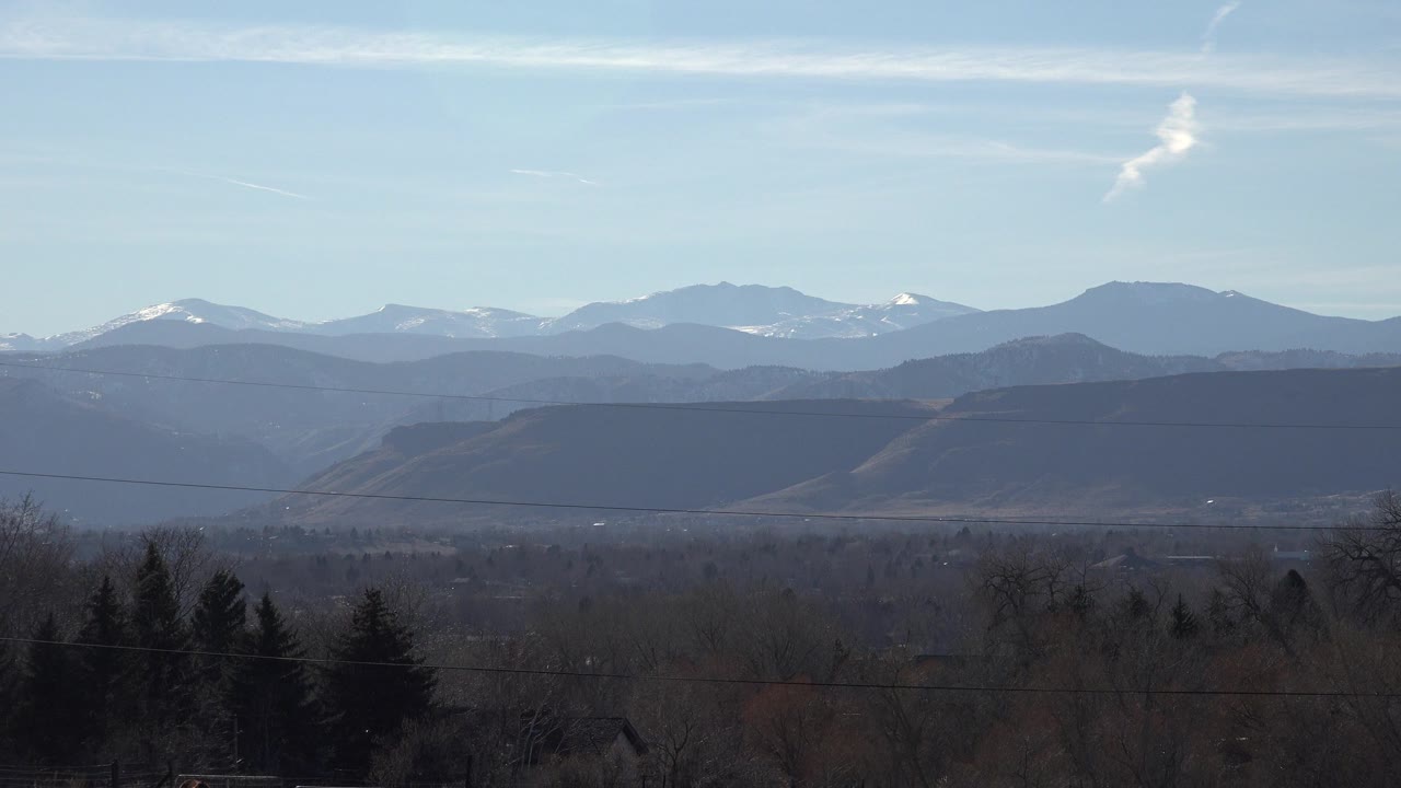 Wide Arvada, Colorado landscape with dark winter trees in the foreground, hazy foothills, distant snow-dusted peaks, and no visible people.