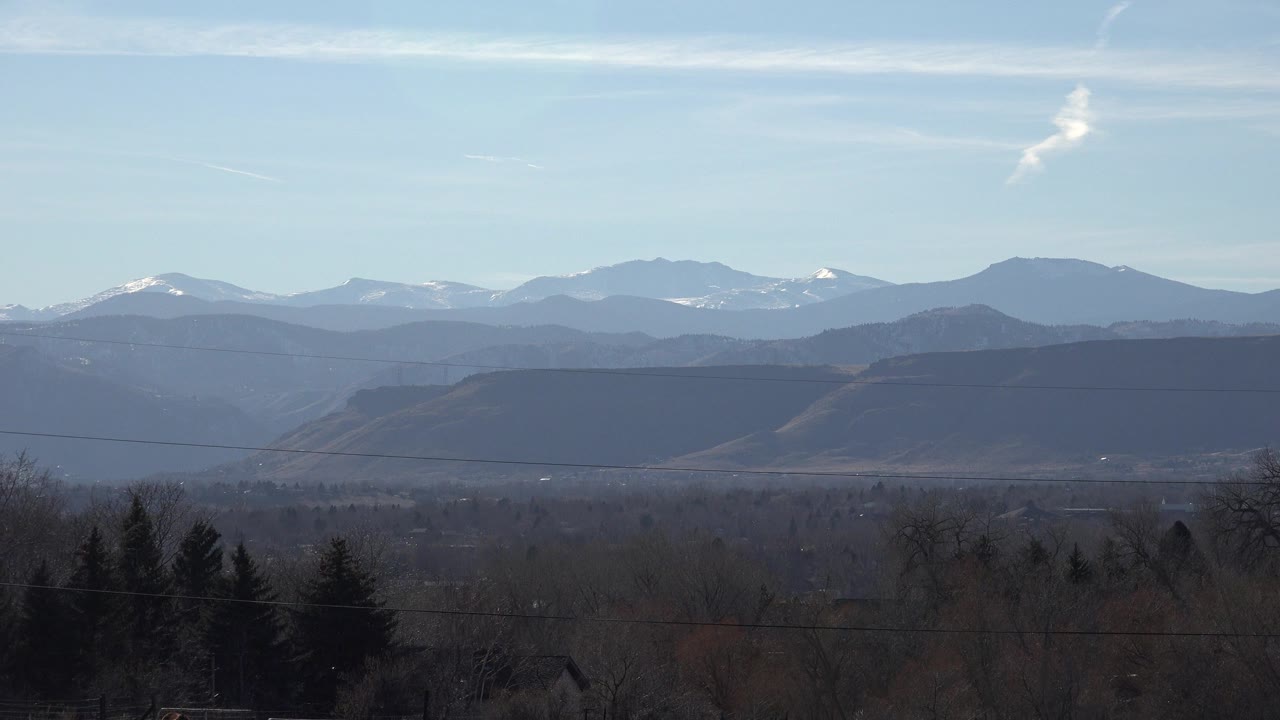Arvada, Colorado foothills view with a flat dark ridge across the midground, faint snowy peaks behind it, foreground trees, and no visible people.