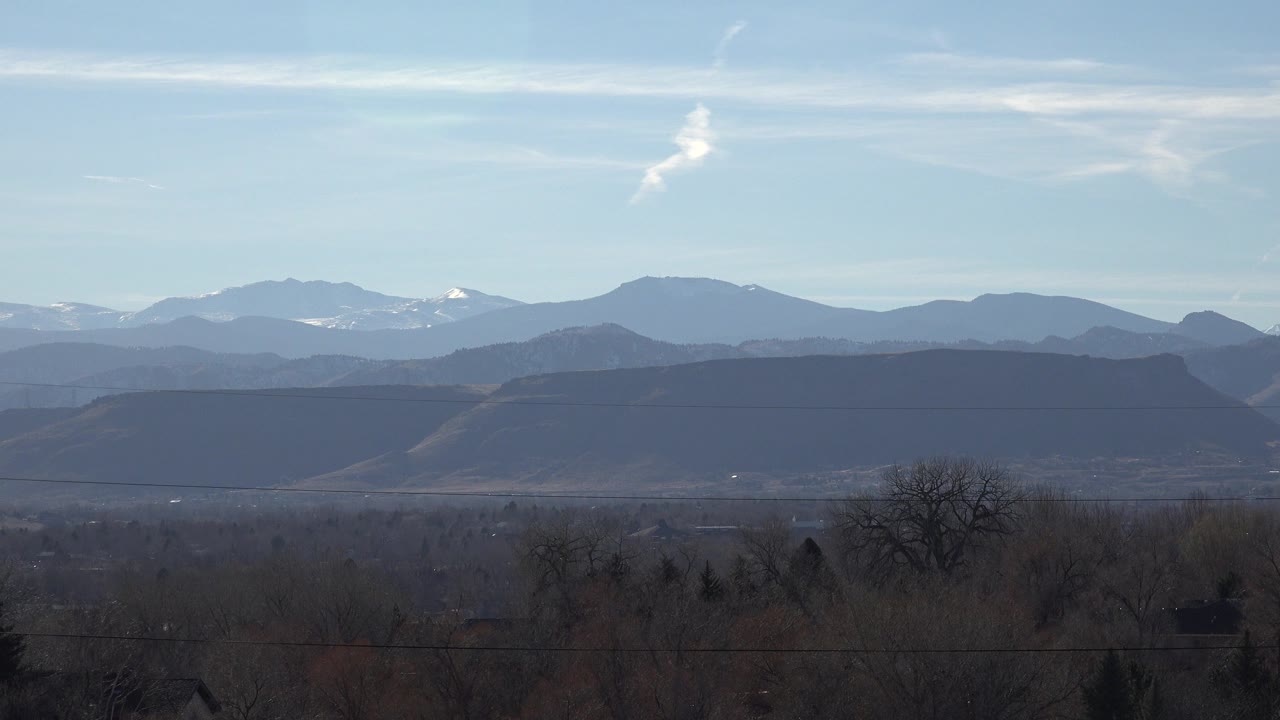Arvada, Colorado mountain ridges appear in hazy layers below pale blue sky, with dark winter trees and no visible people.