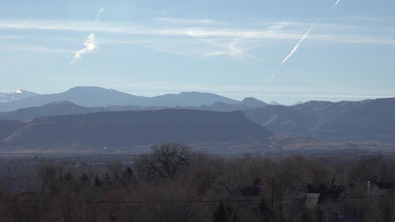 Wide Arvada, Colorado view of layered mountain ridges under pale blue sky, with leafless trees, distant rooftops, a utility line, and no visible people.