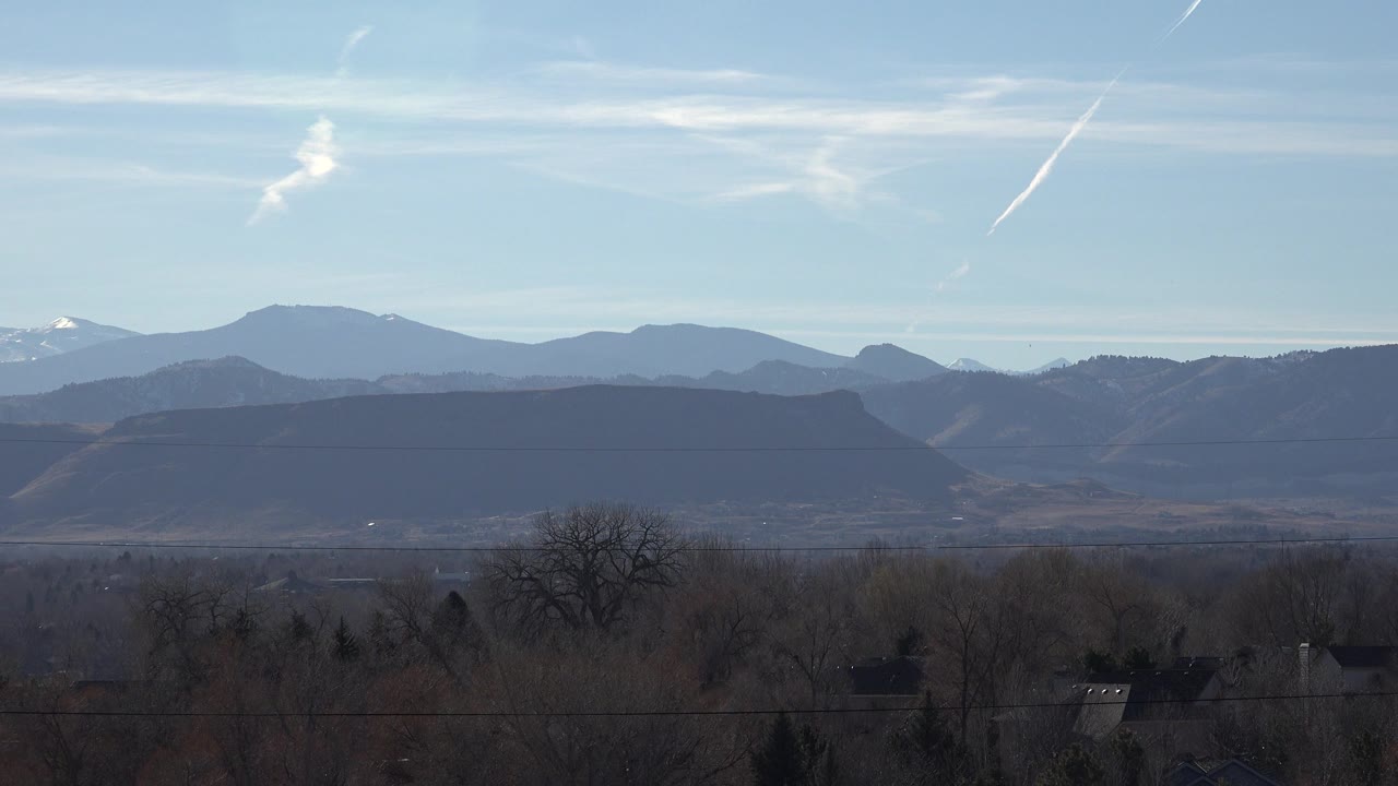 Arvada, Colorado landscape with dark foreground trees and rooftops beneath hazy mountain layers, pale high clouds, and no visible people.