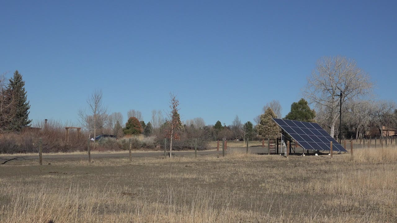 Opening frame of a ground-mounted solar panel in an Arvada, Colorado field with dry grass, leafless trees, fence posts, distant homes, and no visible people.