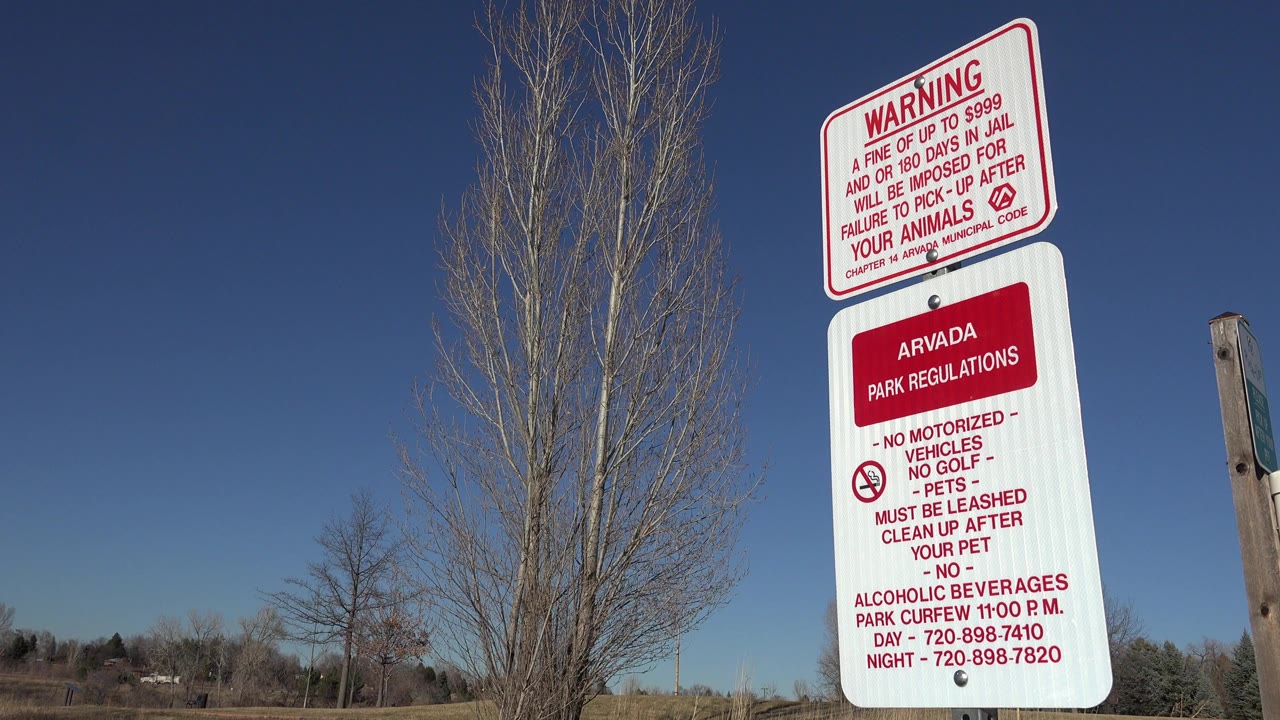 In Arvada, Colorado, a red-lettered park warning and regulations sign stands at the right of frame near a tall leafless tree, with no visible people.