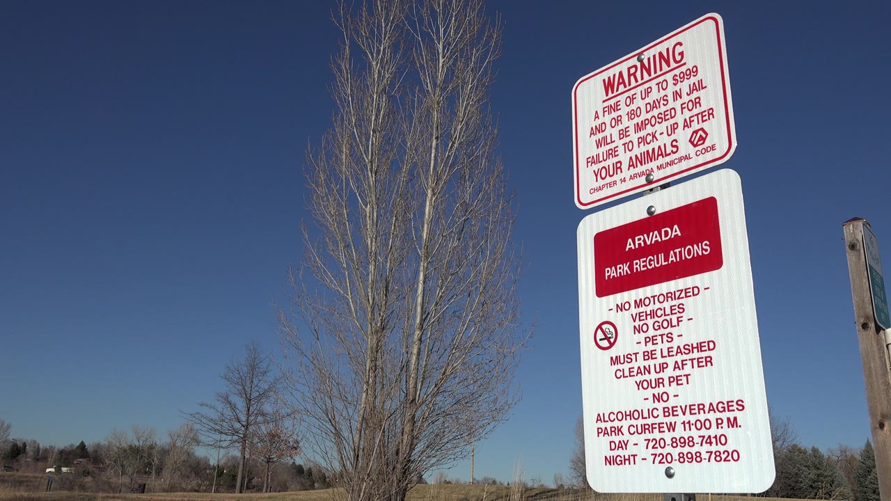 Close view of a white park sign with warning text and leafless trees in the background.